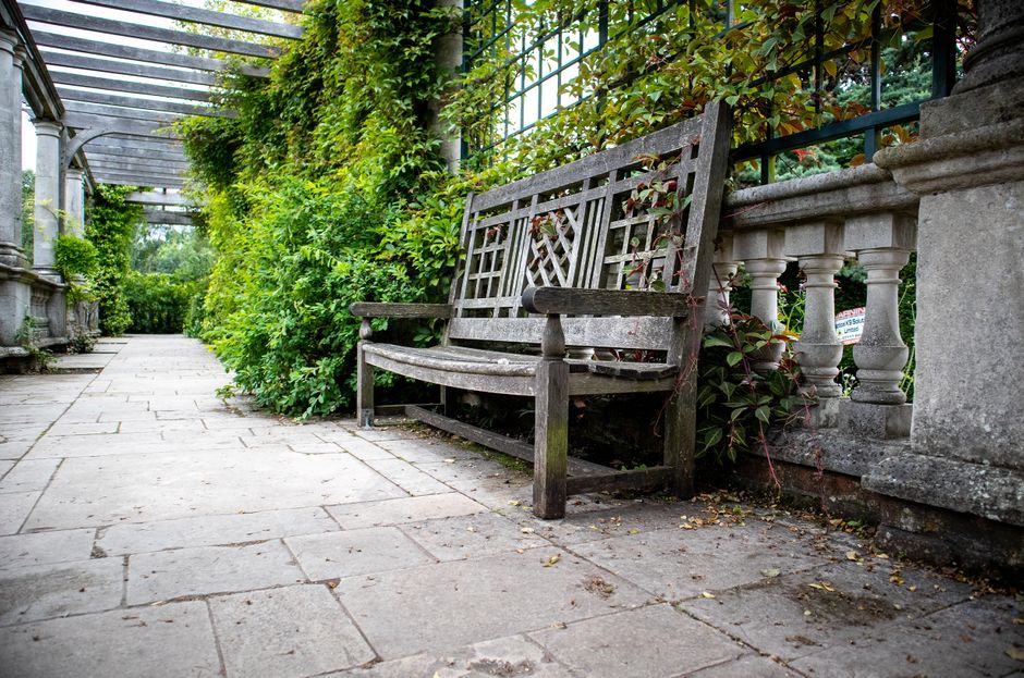 A weathered wooden garden bench with a lattice backrest and armrests positioned on a stone-paved pathway in a well-maintained outdoor space. The bench is placed beside a decorative stone balustrade, b